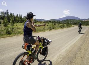 A young woman riding a freehand bicycle aims at the person ahead on a bicycle tour with a slingshot